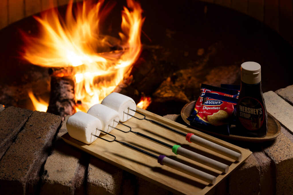 焼きマシュマロの準備（串とビスケット、チョコレートソース）と、その奥に焚き火の炎